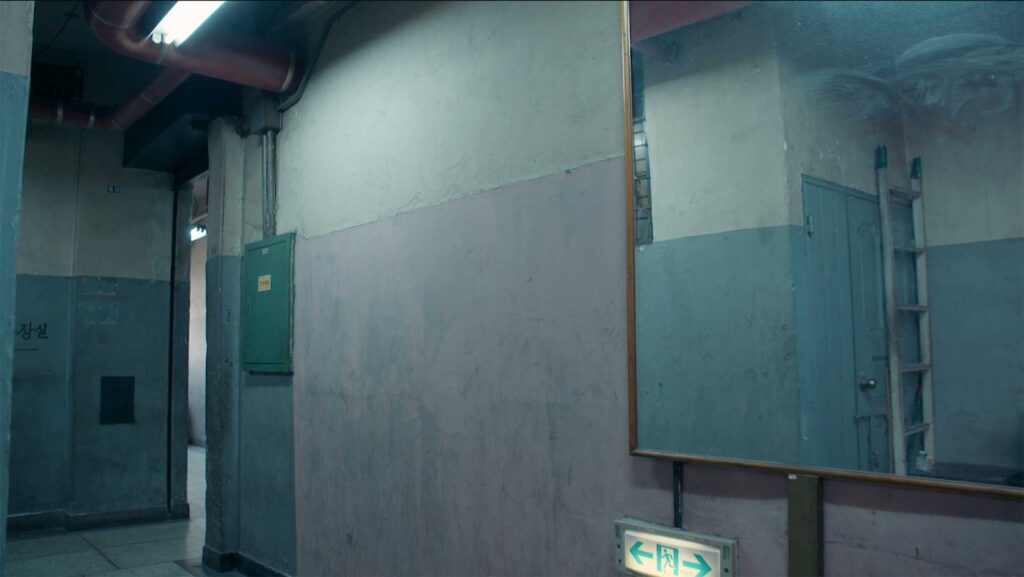 Industrial corridor with worn painted walls, exposed pipes, and mirror reflection suggesting shared living spaces within worker housing from the 1970s