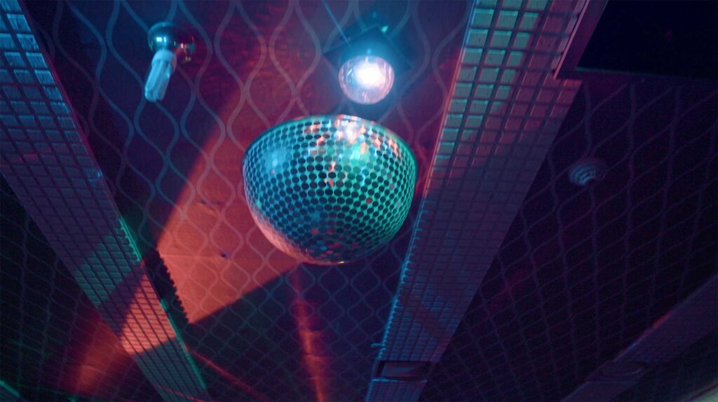 Disco ball suspended beneath patterned ceiling with colored light reflections evoking brief escape and nightlife culture within worker communities