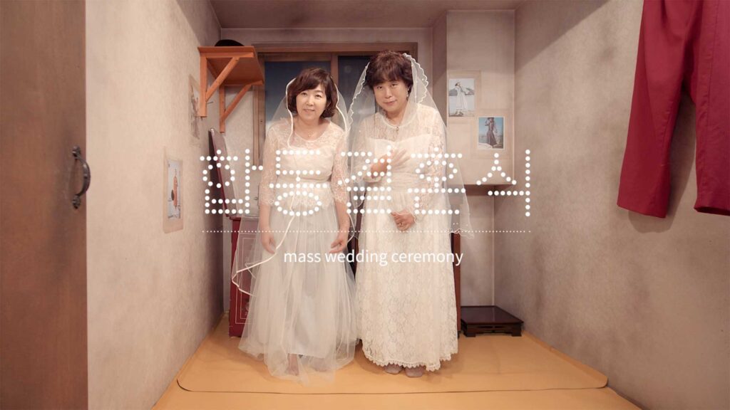 Two women in wedding dresses inside a small domestic room referencing mass wedding ceremonies during Korea’s rapid industrialization period