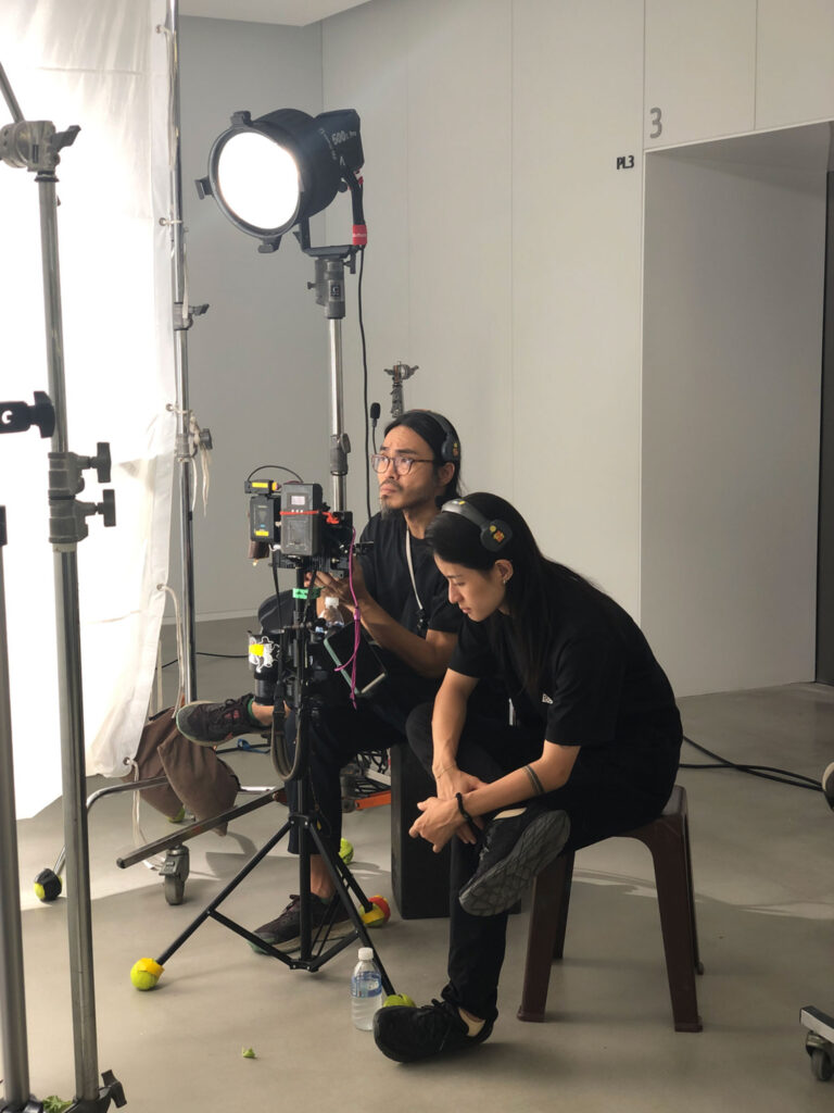 Camera operator and assistant monitoring a studio setup, seated beside lighting equipment while reviewing framing and exposure during a controlled shoot