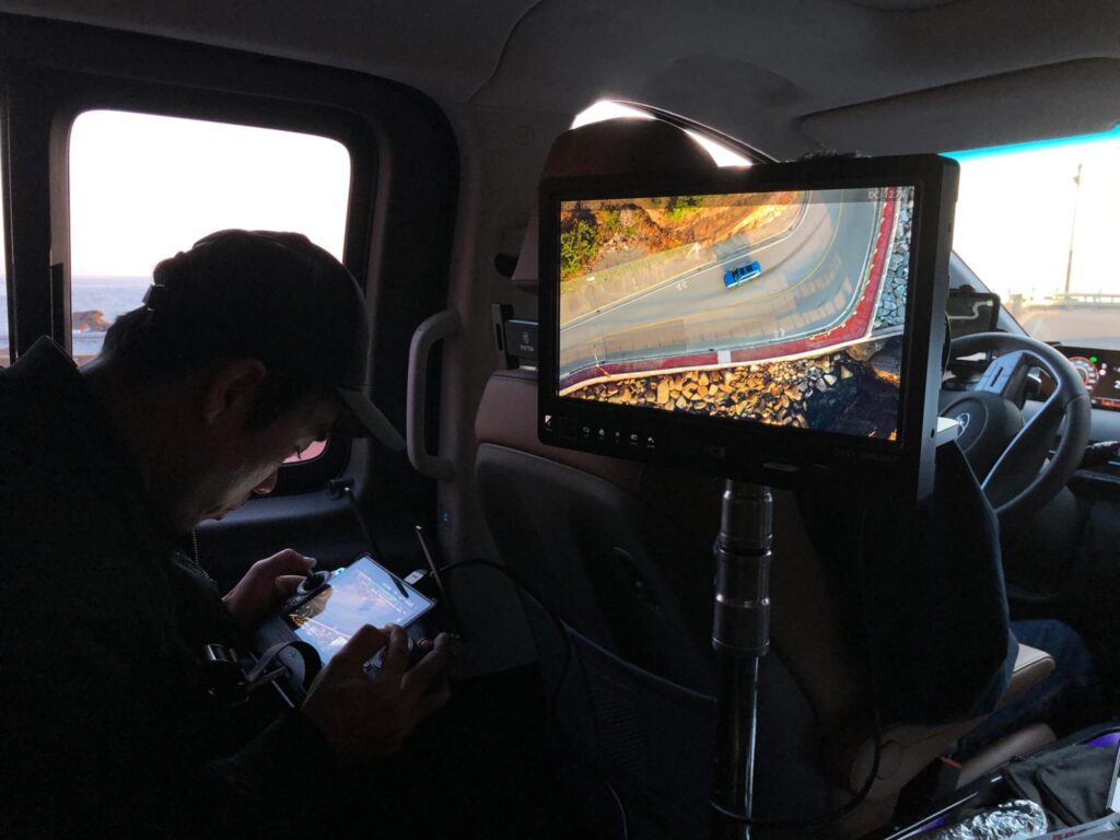 Crew member monitoring drone footage inside a moving vehicle, reviewing an overhead coastal road shot while adjusting camera and flight settings