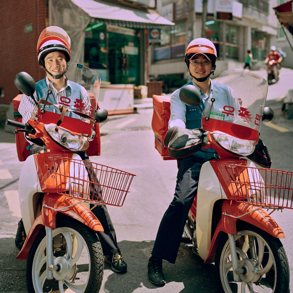 Two Korean postal service delivery drivers, uniformed, sit on their resting motorcycles in a side street in Seoul, South Korea. Helmet visors flipped up