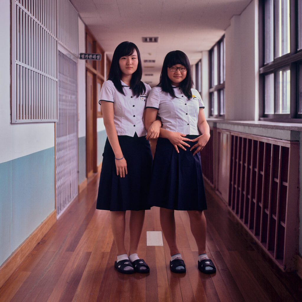 Two female Korean high school students in uniform pose in the main classroom alleyway of a school building, creating a contrast with their slippers and socks, adding a touch of individuality to their ensemble