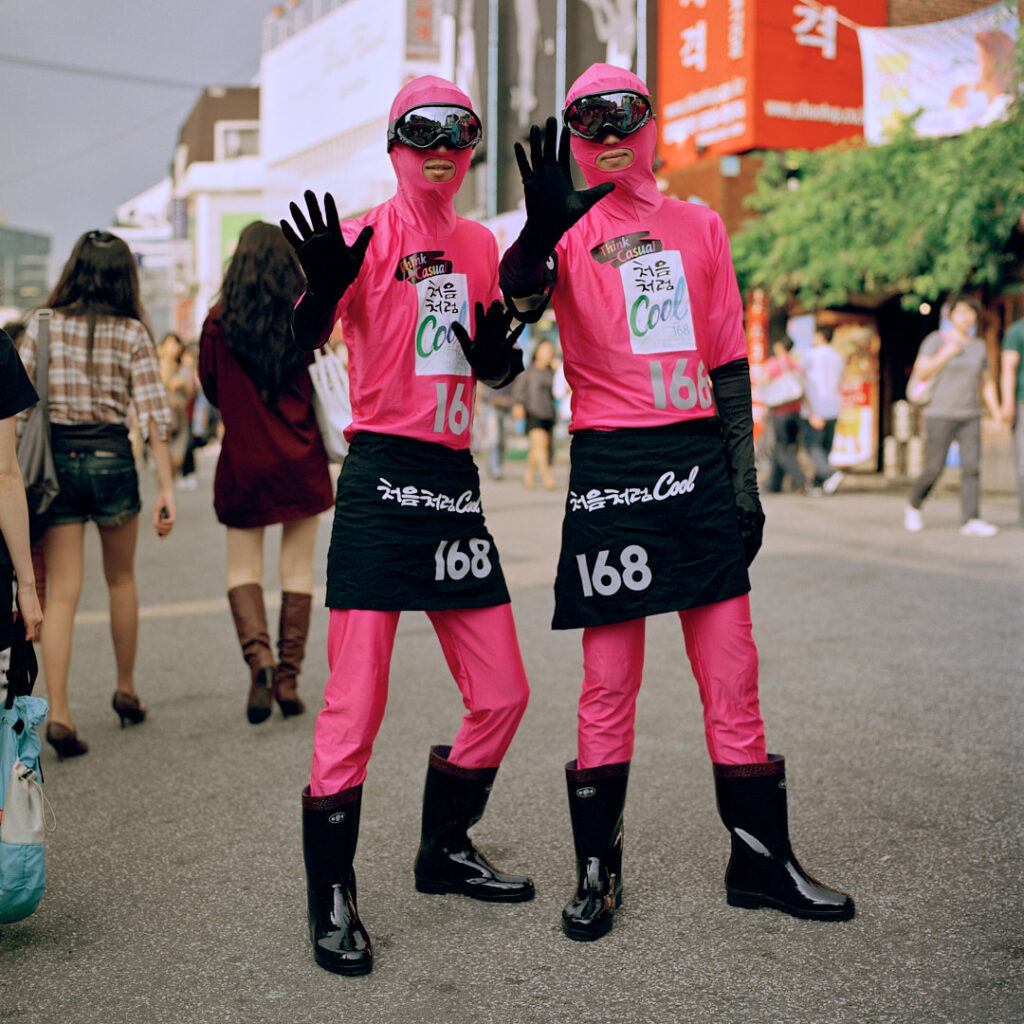 Two males stroll the streets of Seoul in funky, space-themed uniforms to advertise a product. Clad in pink pants, tight space hoodies, black dresses, gum boots, and ski visors, they grab attention