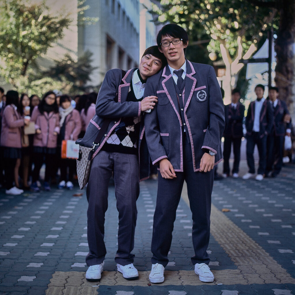 Male high school students in uniform and white sneakers pose, one resting his head on the other's shoulder, amidst a group of classmates